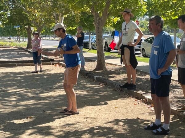 A group of people standing outdoors under the shade of trees, with some looking at the man in the foreground who is playing shuffleboard on a dirt court.