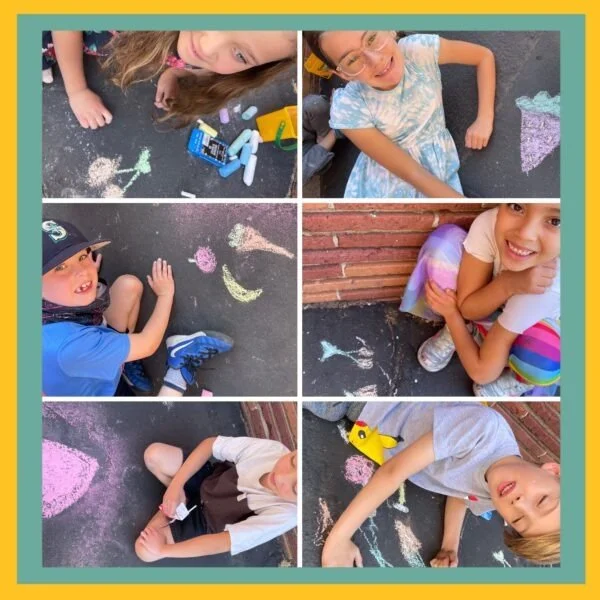 Six children drawing with sidewalk chalk on asphalt, some sitting or lying down, with colorful chalk drawings of hearts, flowers, and animals.