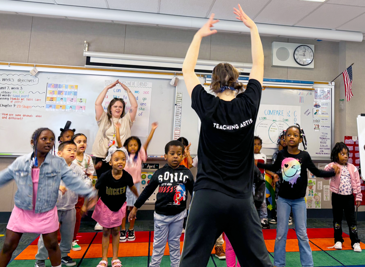 A classroom with young children participating in a dance or movement activity, led by an instructor wearing a black shirt labeled 'Teaching Artist'. There are whiteboards and educational posters on the walls.