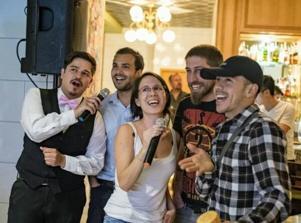 Group of five friends smiling and singing karaoke in a bar, with microphones in hand.