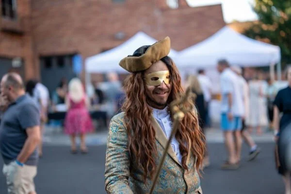 Person dressed as a masked pirate with long curly hair and a colorful coat at an outdoor event with tents and people in the background.
