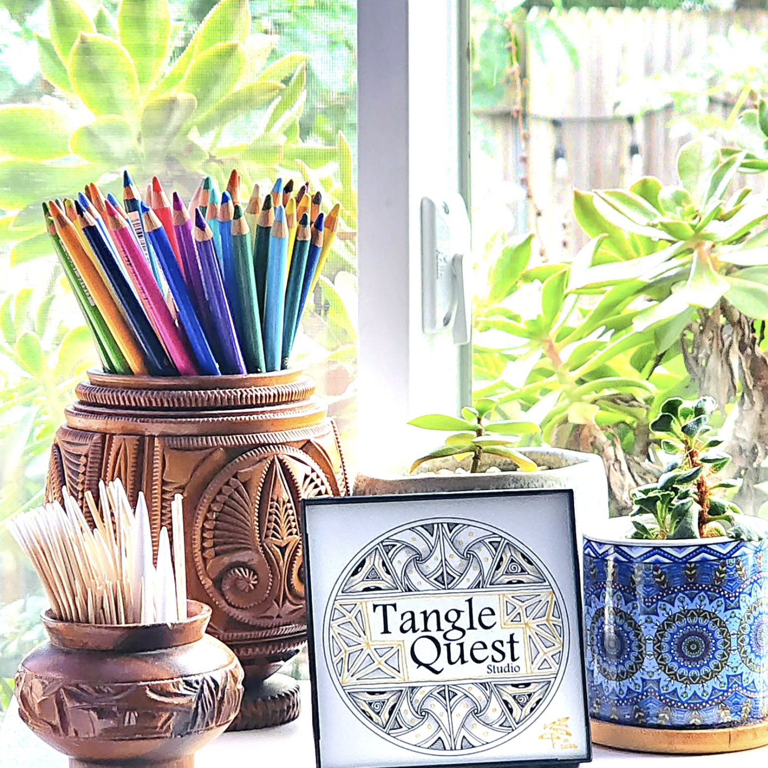 Colorful colored pencils in a wooden holder on a windowsill with plants outside, a sign reading 'Tangle Quest Studio,' and decorated pots with plants.