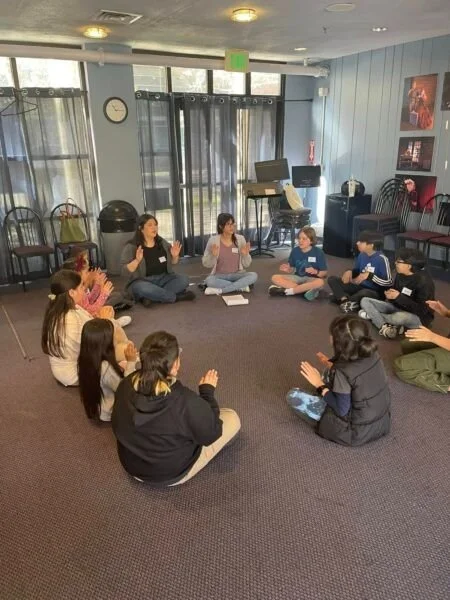 Children sitting in a circle on the carpet in a room, participating in a group activity or discussion with two adults leading the session.
