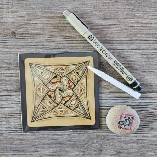 A decorative square-shaped object with intricate geometric and swirling patterns, a white marker pen labeled 'MICRON 01', and a round white object with red and blue markings on a wooden surface.