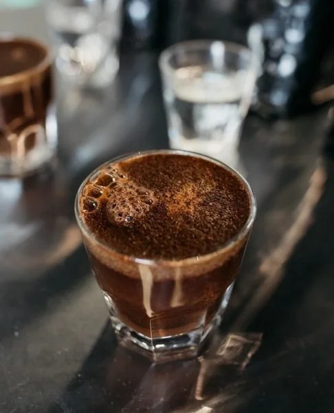 Close-up of a glass of hot chocolate or coffee on a dark table with a glass of water and other mugs or cups in the background.