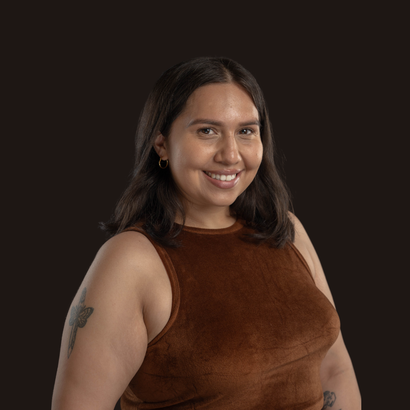 A young woman with shoulder-length dark hair, wearing a brown sleeveless top and small hoop earrings, smiling at the camera against a dark background.