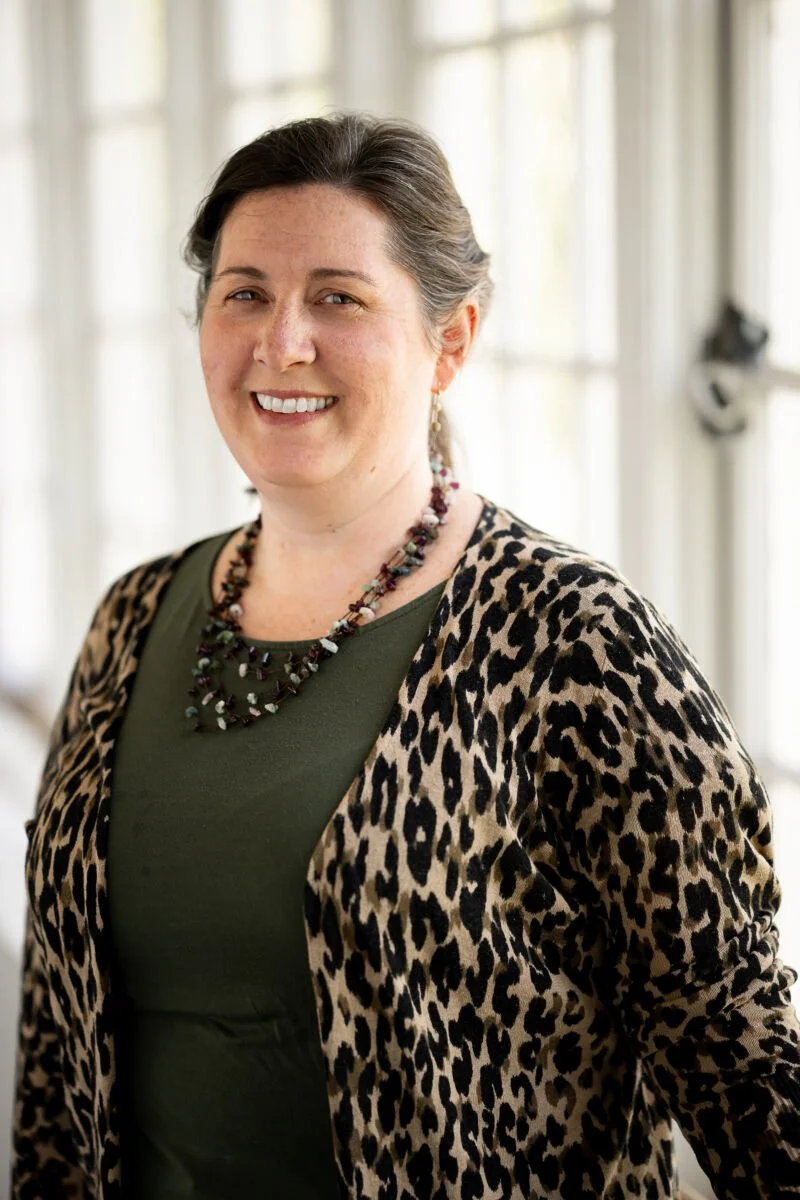 A woman with brown hair, smiling and wearing a leopard print jacket over an olive green top, with a beaded necklace, standing in a bright room with sheer curtains.