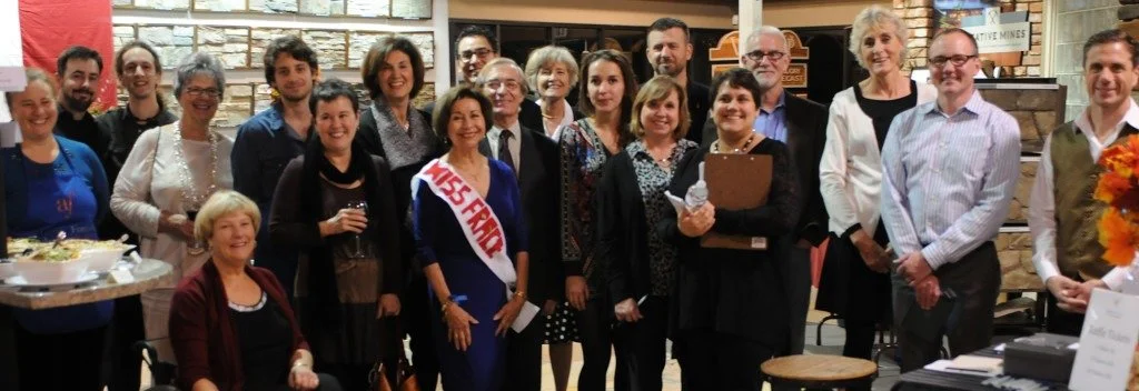 Group of people gathered inside a restaurant or store, smiling for a photo. Some are holding drinks or food, and there is a woman in the center wearing a sash that says 'MISS FRANCE.'