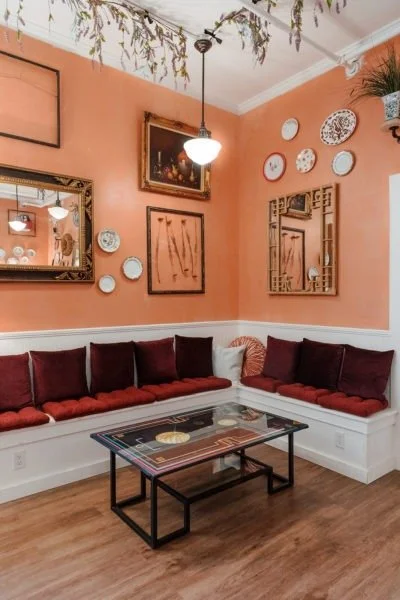 Living room with peach-colored walls, decorated with various framed pictures and decorative plates, featuring a white built-in bench with red and white cushions, a glass coffee table, a hanging light fixture, and potted plants on the ceiling and wall shelves.