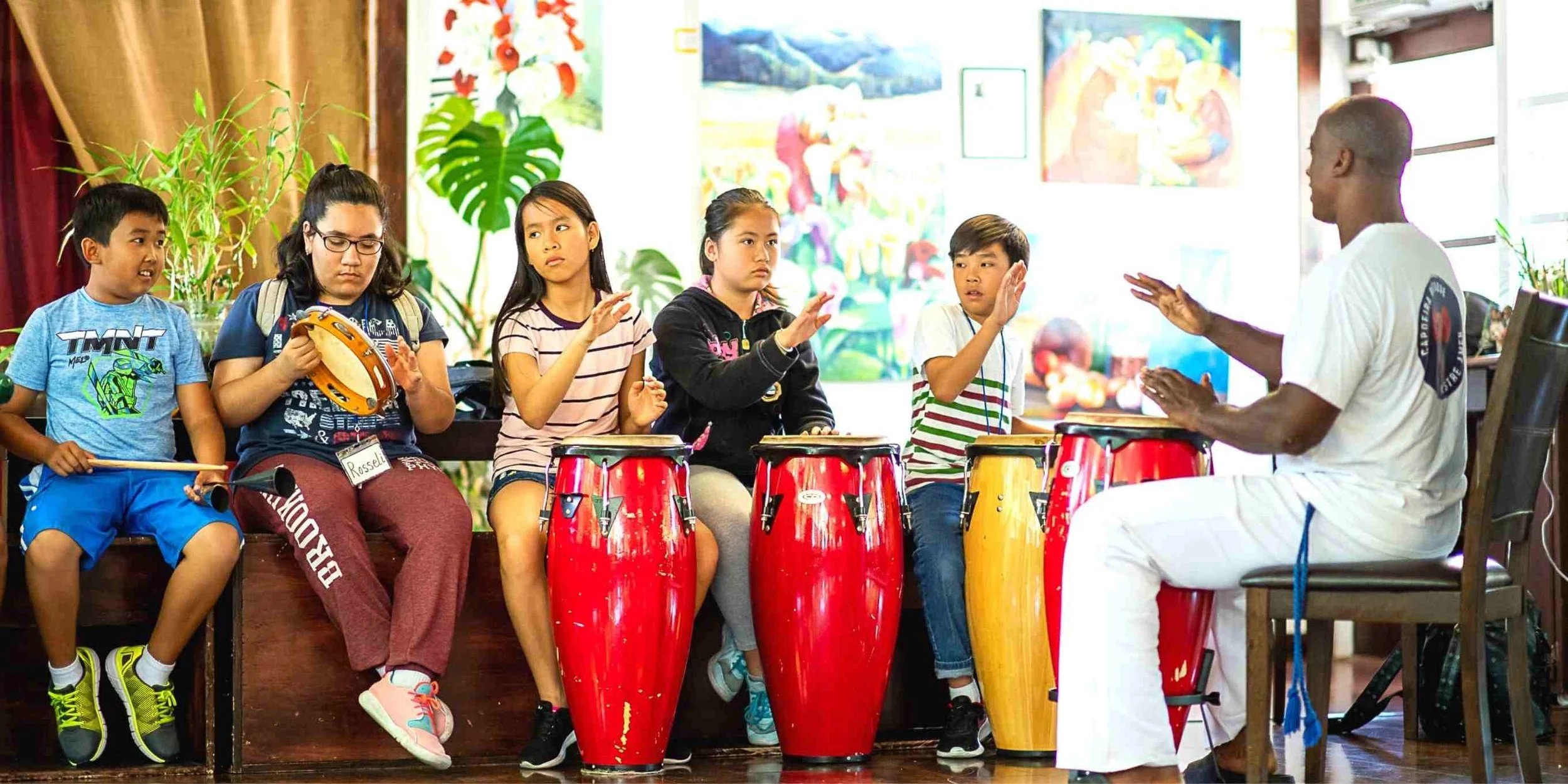 Children sitting and playing drums during a music lesson or performance, led by an instructor.