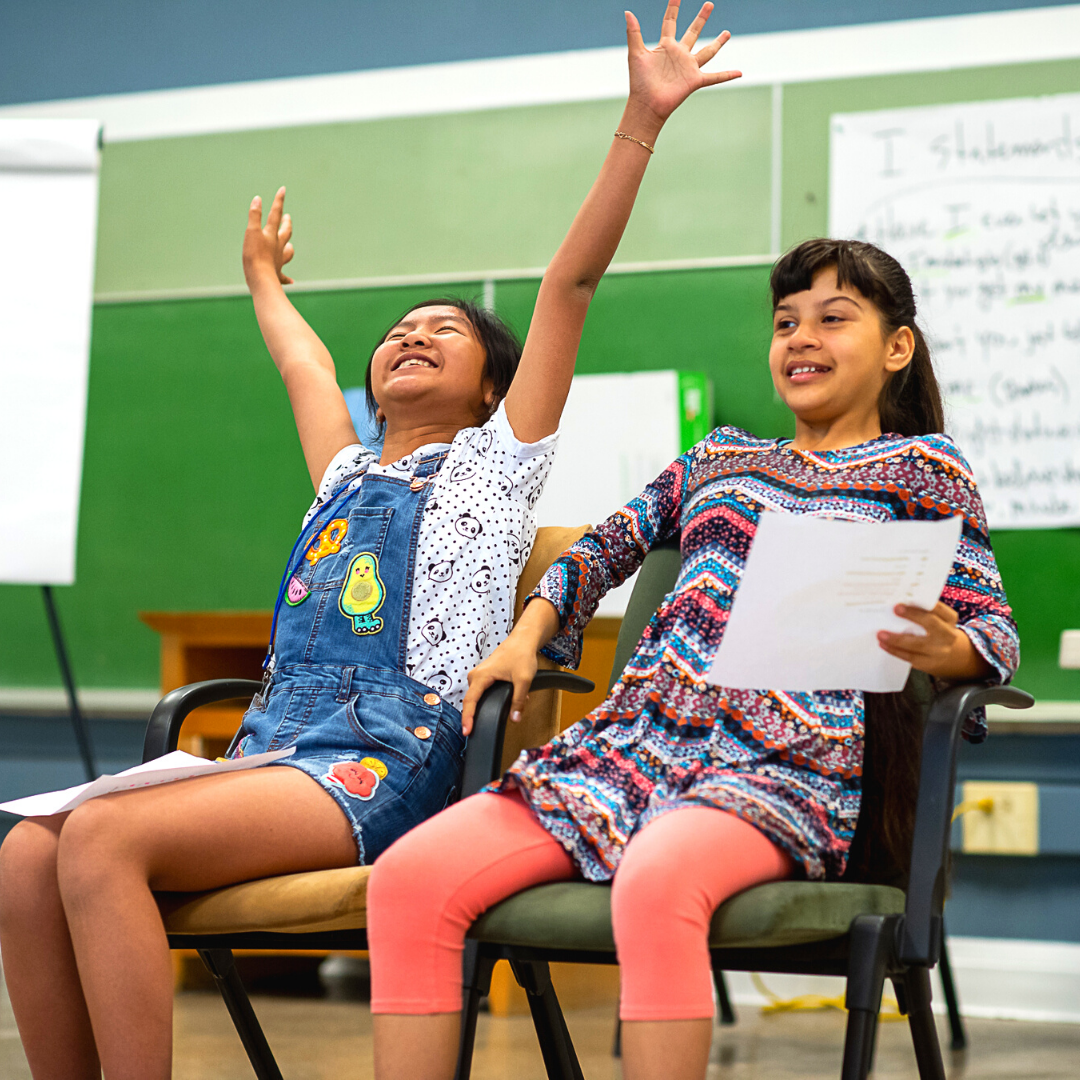 Two young girls sitting in chairs in a classroom, one raising her hand with a big smile, the other holding papers, with a green chalkboard and writing in the background.