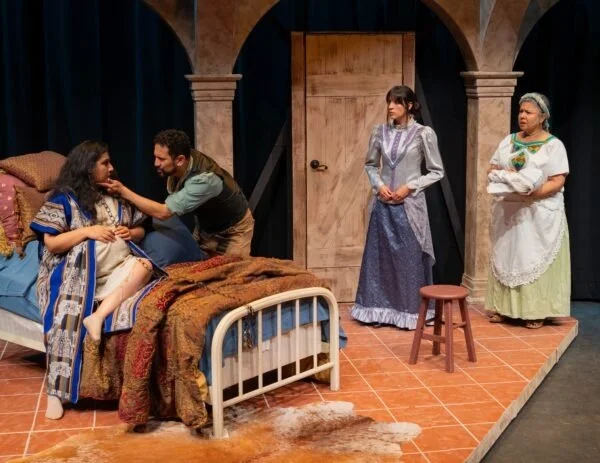 Three women and one man on a stage set, with two women sitting on a bed, one woman standing with arms folded, and one woman standing holding a pillow, in a scene from a play.