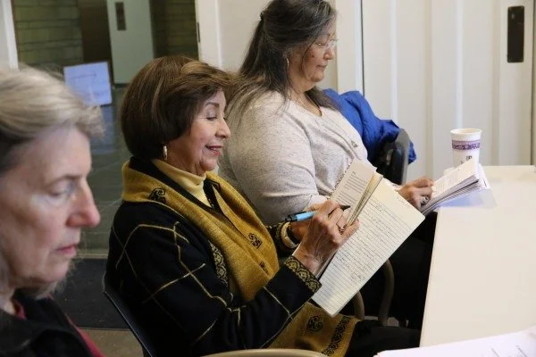 Three women sitting at a table, reading books or pamphlets, in an indoor setting.