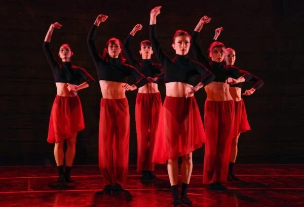 Six female dancers in black crop tops and red skirts perform a synchronized dance on stage, raising their arms in a choreographed pose.