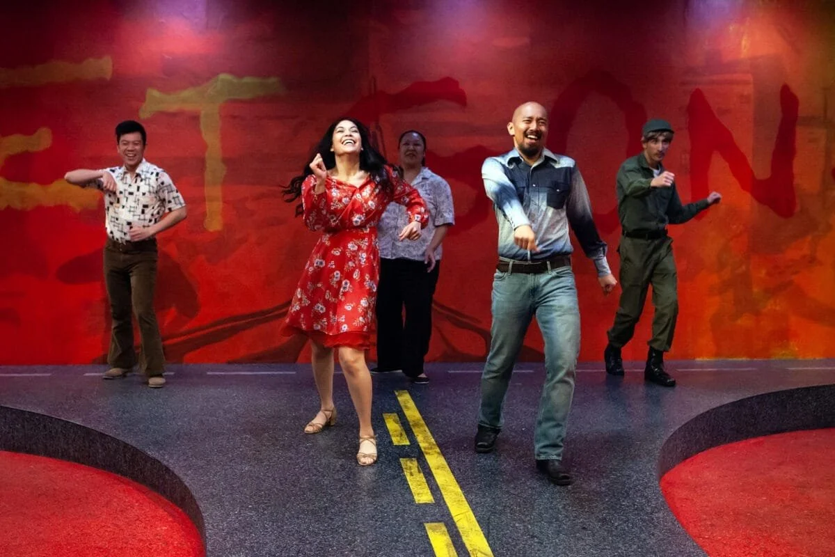 Group of five people on stage dancing and smiling, with a vibrant red and orange background.