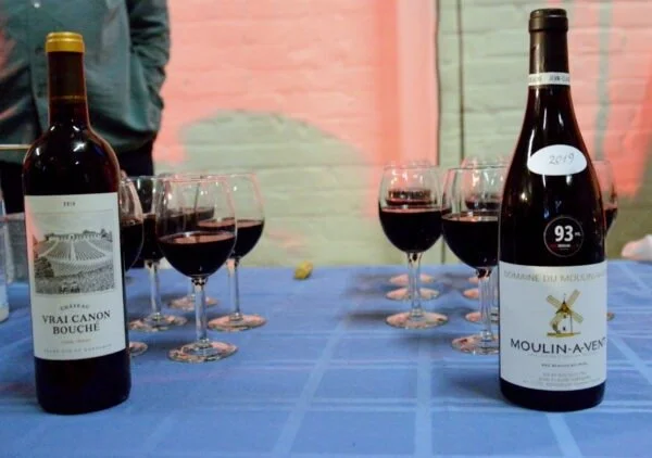Six glasses of red wine on a blue tablecloth, with two bottles of red wine in the foreground, set against a colorful wall.