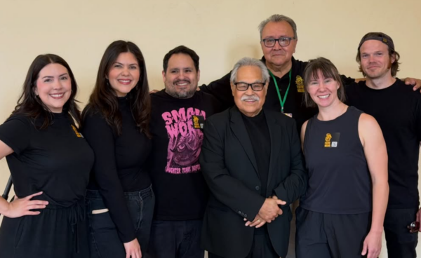 Six people standing side by side, smiling, in front of a beige wall, with some wearing black T-shirts and one person in a dark blazer and glasses.