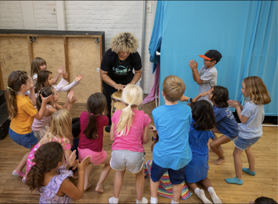 Group of children participating in a drum circle led by an instructor, in a room with a brick wall and a blue curtain.
