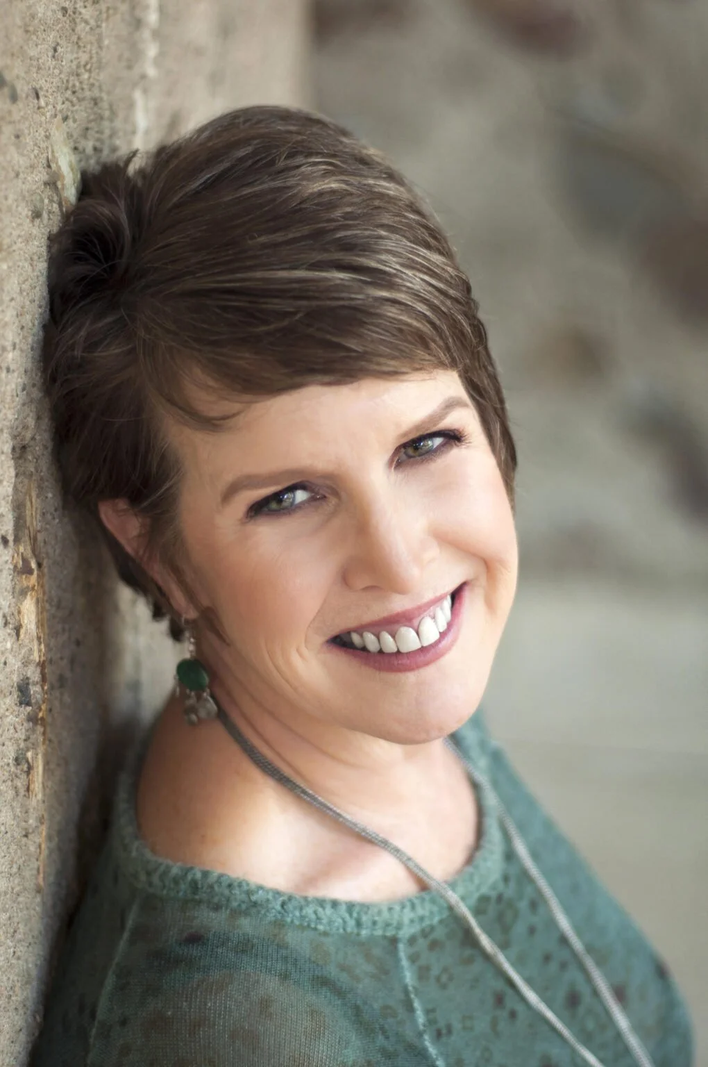 A woman with short brown hair and blue eyes smiling, leaning against a stone wall.