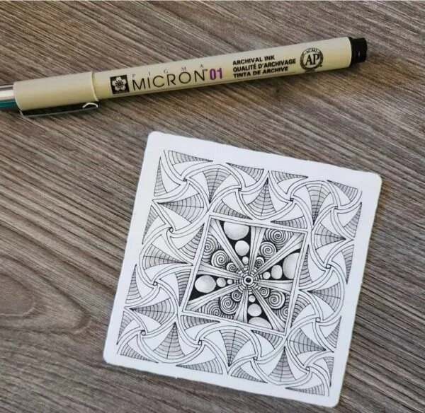 A black and white geometric pattern drawing on a piece of paper and a beige Uni-Ball Micron 01 archival ink pen placed on a wooden surface.