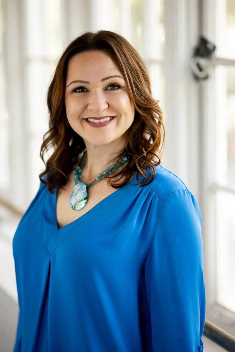 A woman with shoulder-length wavy brown hair, wearing a bright blue blouse and a turquoise necklace with a large pendant, smiling in a room with large windows and sheer curtains.
