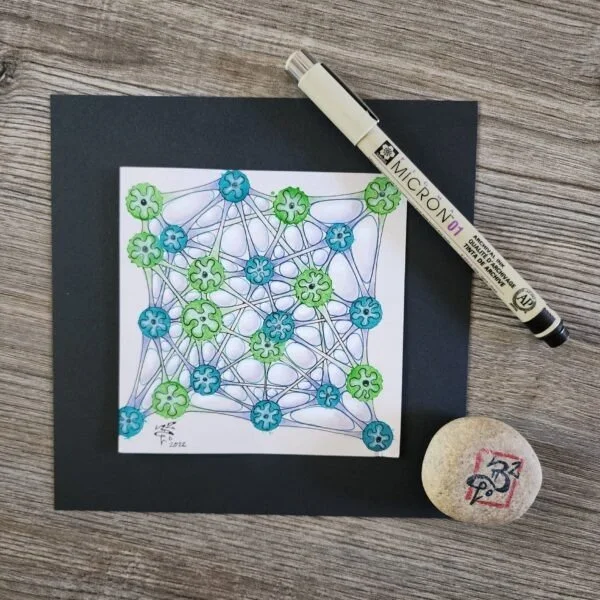 Colorful hand-drawn diagram with interconnected nodes in green and blue, a black marker pen labeled 'MICRON 01', a black sheet, and a small round stone with a red and black drawing, on a wooden surface.