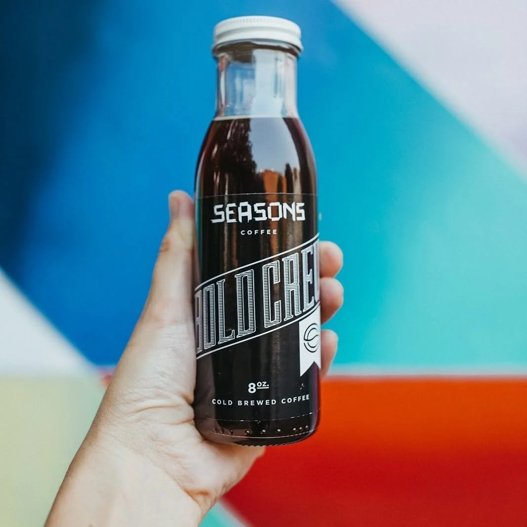 Hand holding a small bottle of Seasons Cold Brew Coffee against a colorful abstract background.