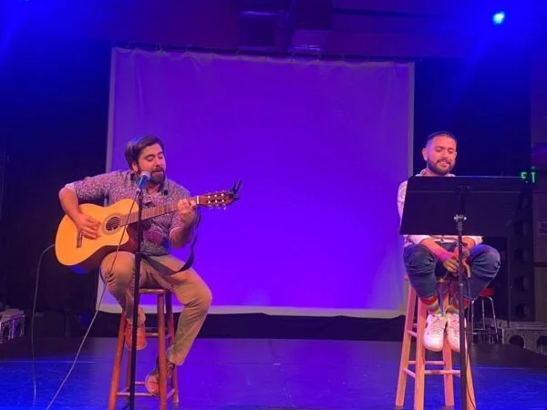 Two men performing on stage with one playing a guitar and the other sitting with a microphone stand in front of him. The background is illuminated with purple lighting.