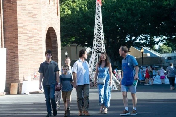 Group of six people, including children and adults, walking outdoors near a brick building and a decorative white tower, with trees and outdoor event booths in the background.