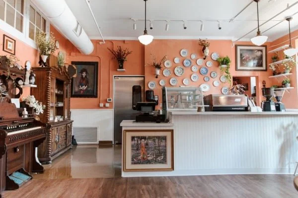 Interior of a cozy cafe with a white counter, decorative plates on the wall, potted plants, and vintage furniture, including a piano and framed artwork.