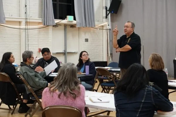 A man standing and speaking with raised finger to a group of people seated around tables in a classroom or meeting room.