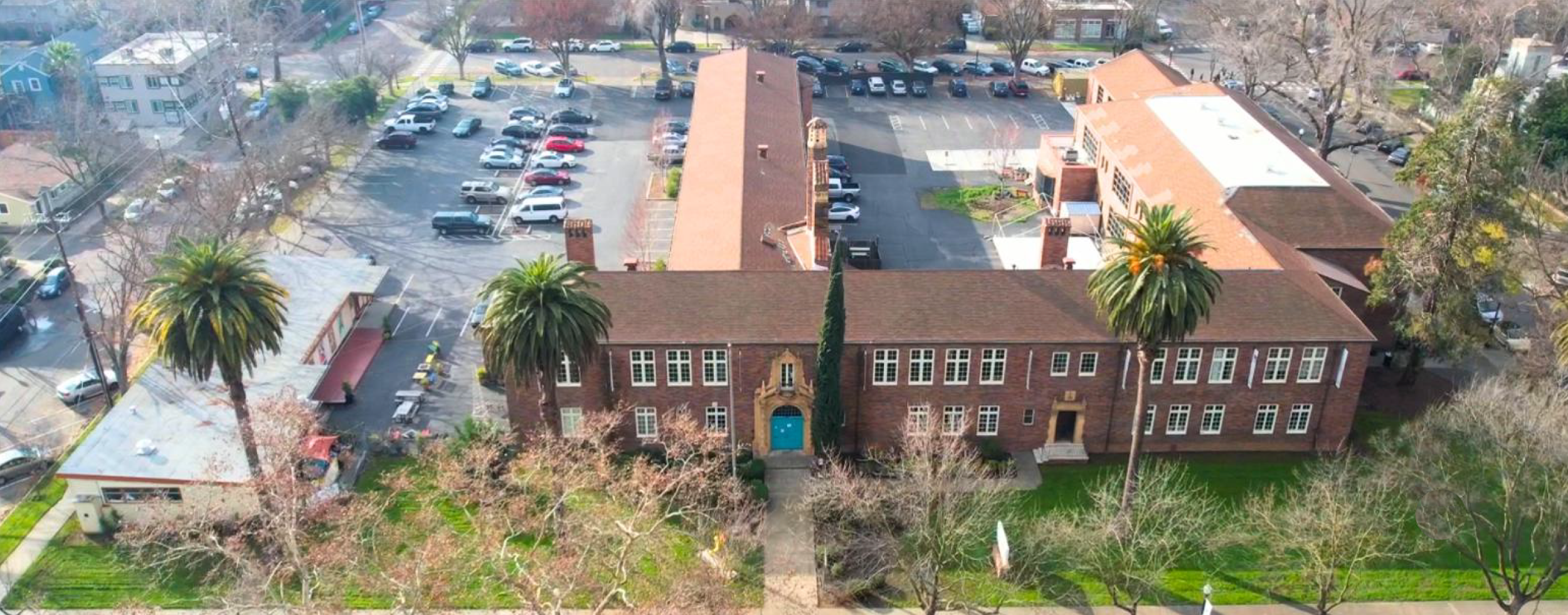 Aerial view of a large brick building with palm trees and a parking lot filled with cars.
