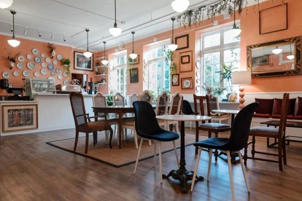 Interior of a cozy cafe with wooden flooring, pastel peach walls decorated with plates and framed art, large windows with green plants, and a variety of tables and chairs.