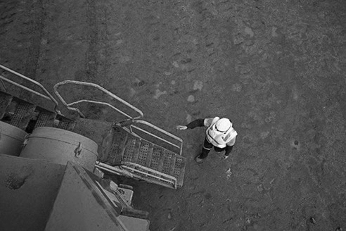 A person in a hoodie and dark clothing stands on a dirt ground near metal stairs and railings, looking upward, seen from a high-angle, black-and-white photograph.