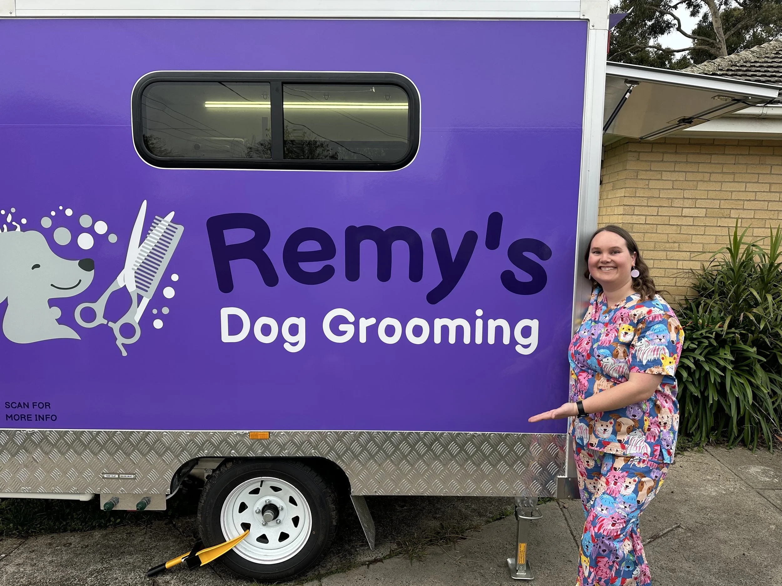 A woman in colorful scrubs standing in front of a purple mobile dog grooming trailer, smiling and pointing at it.