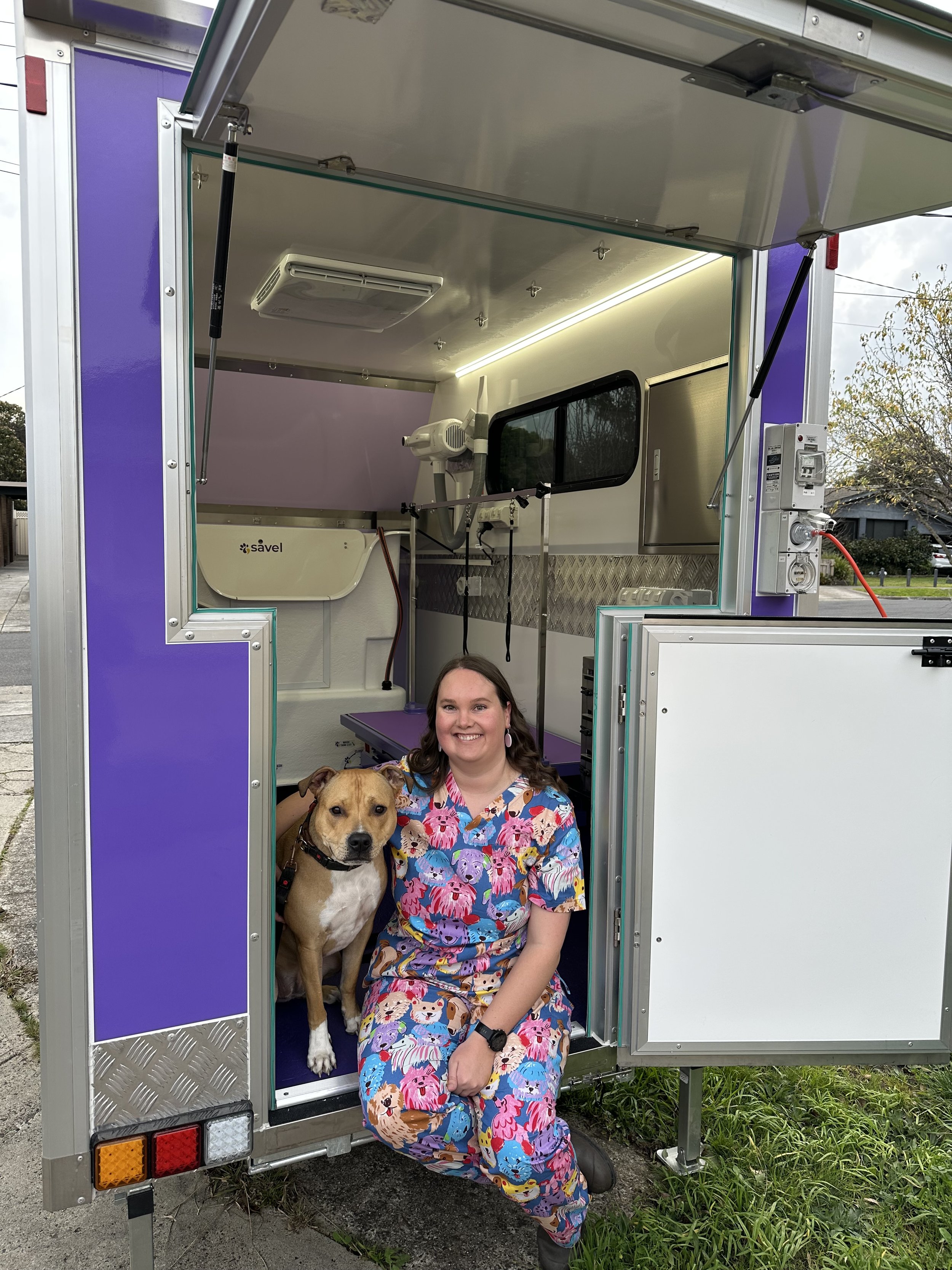 A woman in colorful dog-print scrubs sitting on the ground, smiling, with a dog sitting beside her inside a purple and silver animal rescue trailer, outdoors on grass.