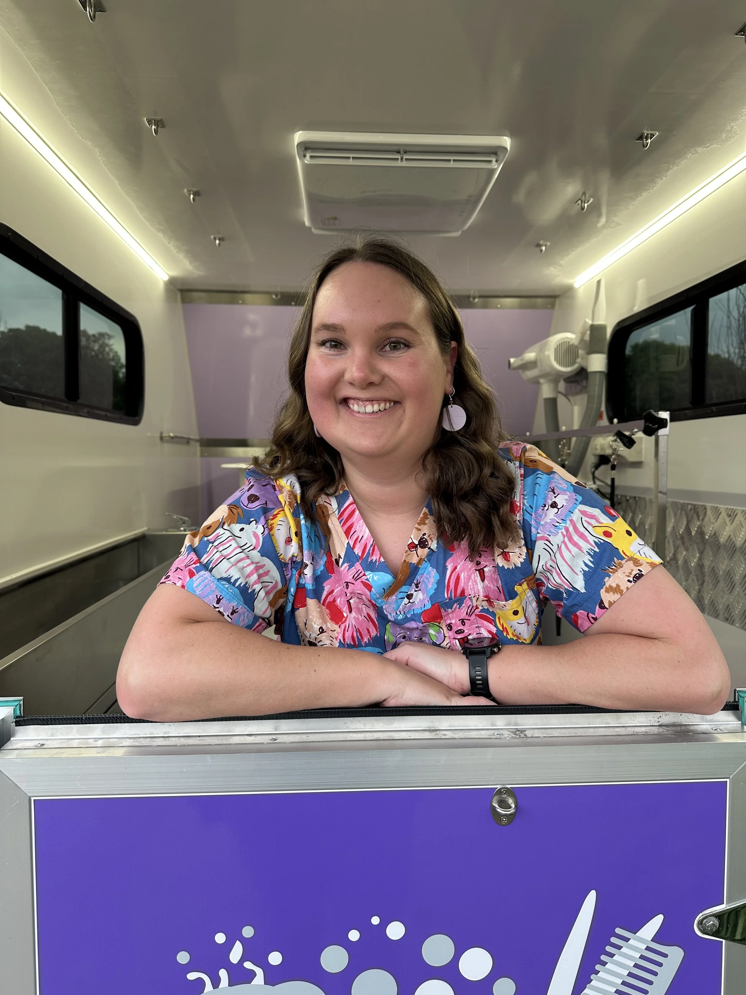 A woman with long, wavy brown hair smiling, sitting behind a counter in a food truck, wearing colorful scrubs with various cartoon characters on them, and a black wristwatch.