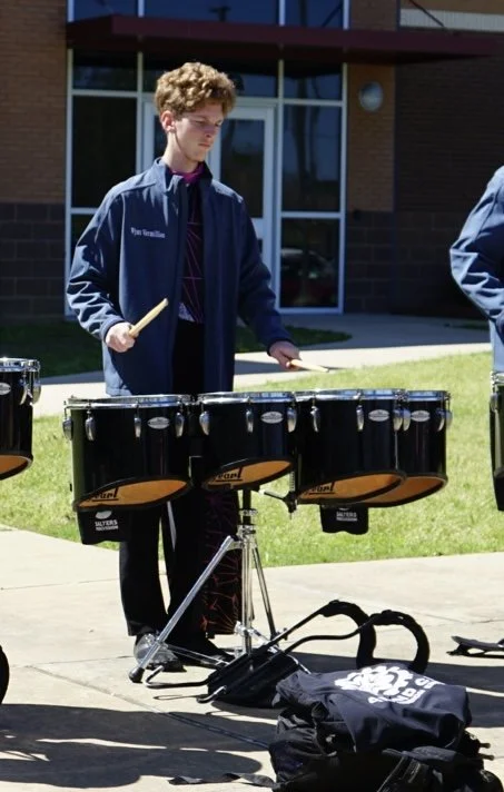 A young man standing outside playing a black percussion instrument on a stand, with a building and sidewalk in the background. He is wearing a dark jacket and holding drumsticks.