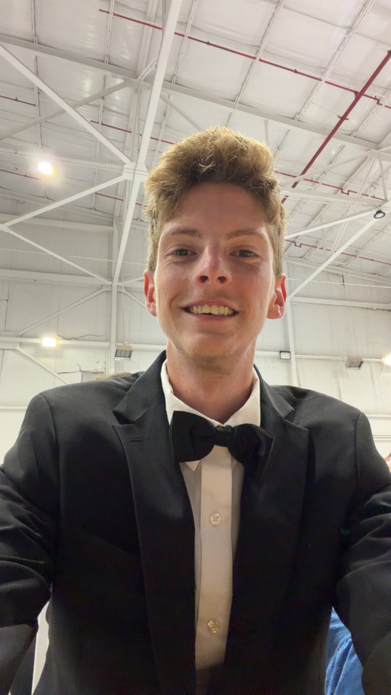 A young man with short, wavy hair taking a selfie. He is wearing a black tuxedo with a white dress shirt and a black bow tie. The background appears to be an indoor space with a high ceiling and visible metal beams and lighting.
