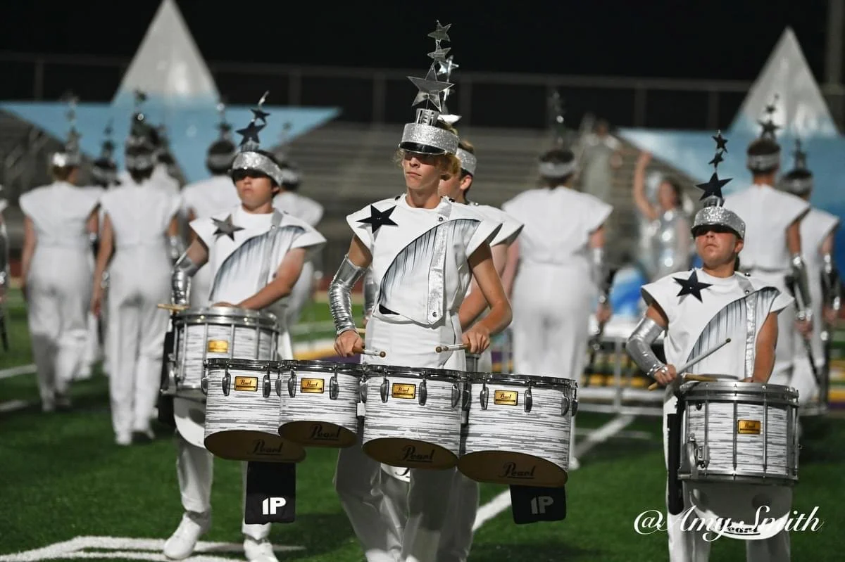 Band members dressed in white costumes with star and silver hat decorations, playing drums on a football field at night.