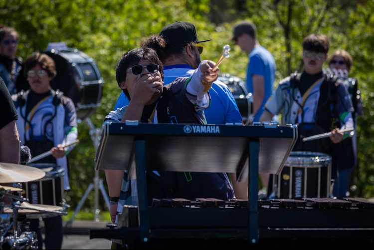Music students practicing with percussion instruments outdoors, including a keyboard and drums, on a sunny day.