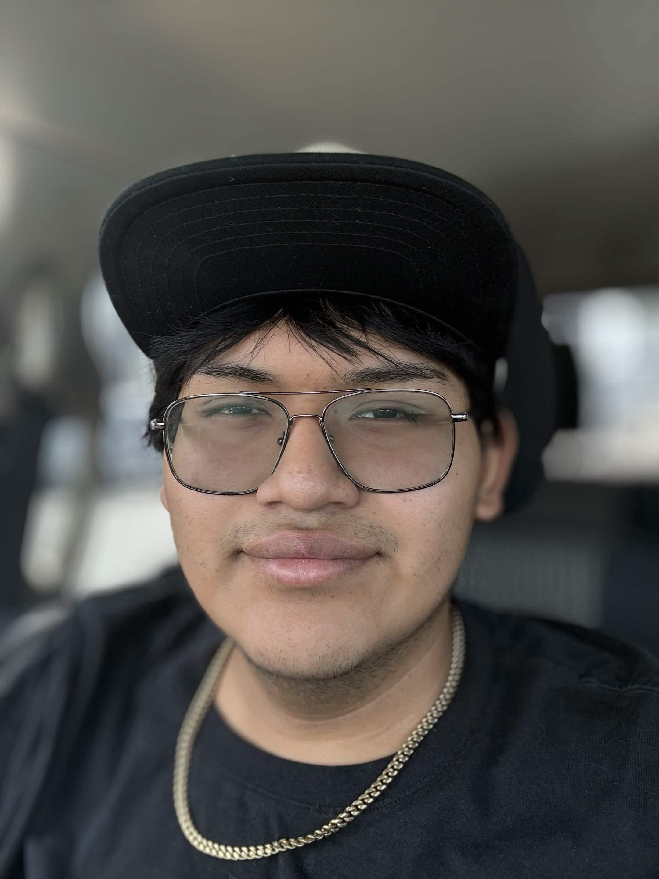 A young man with glasses and wearing a black baseball cap smiling inside a vehicle, with a silver chain around his neck.