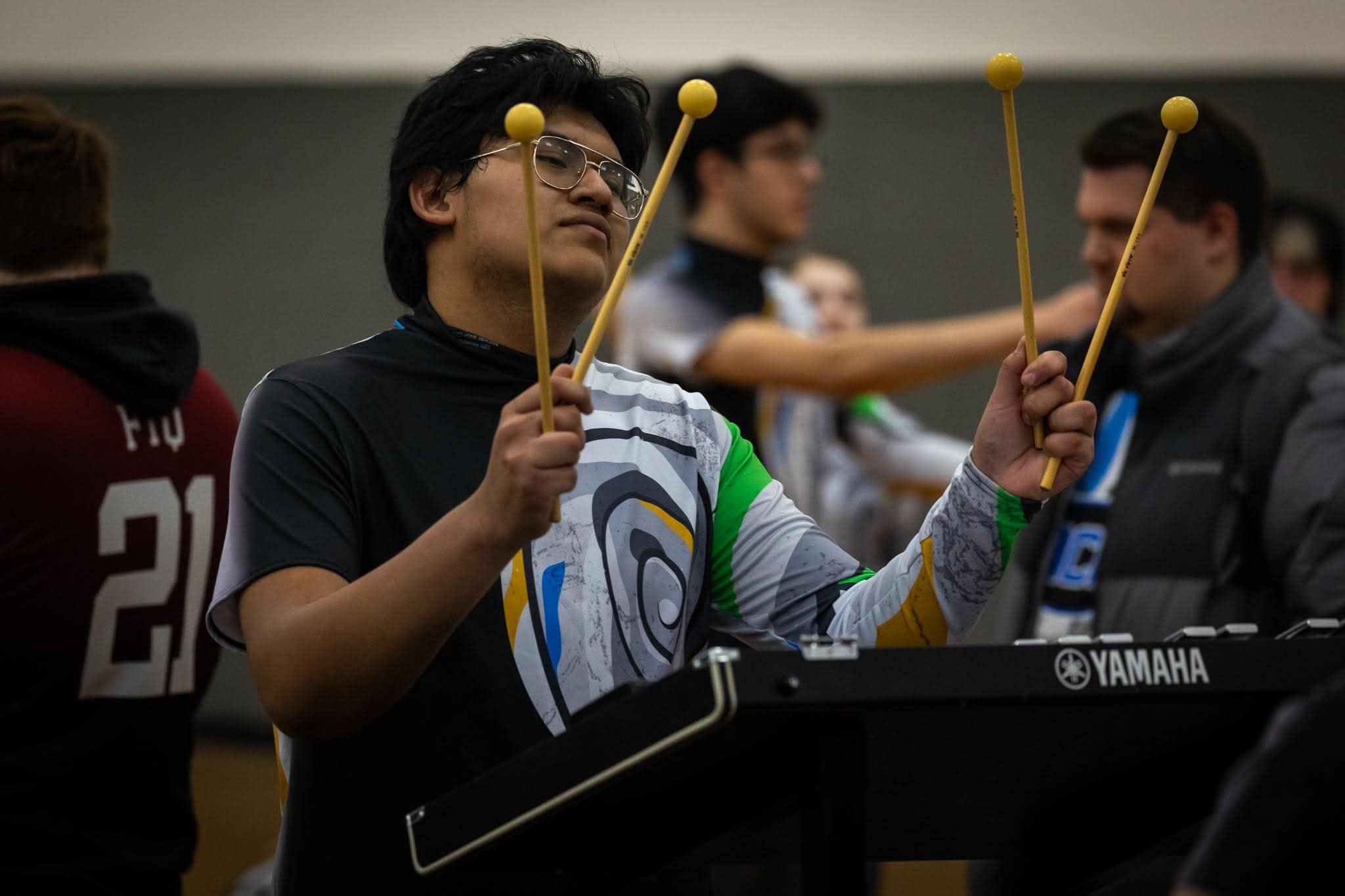 Young man with glasses and a patterned sports shirt holding drumsticks in a group setting, possibly a music room or rehearsal space.