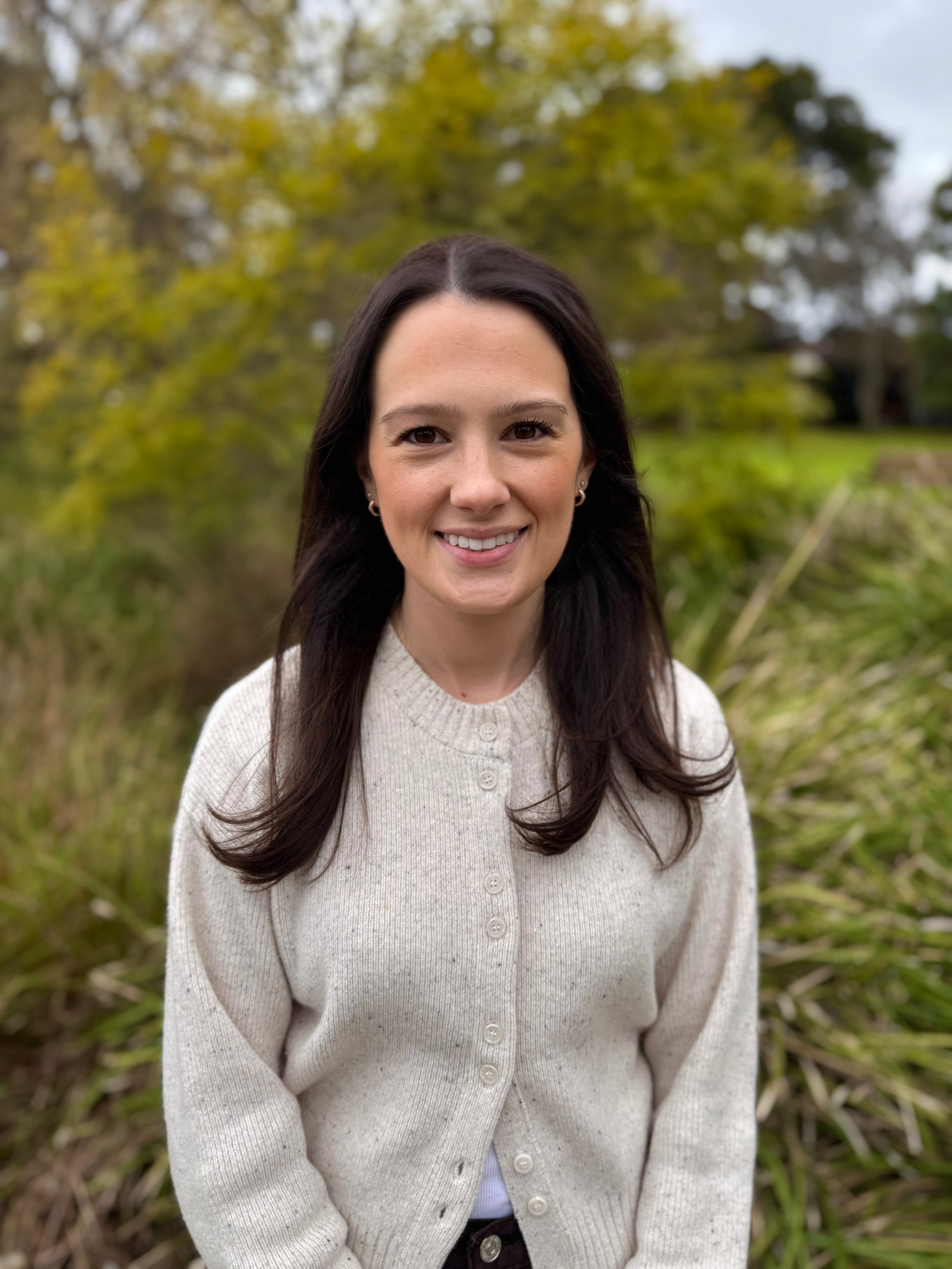 A woman with long dark hair, brown eyes, and earrings smiling outdoors in front of green leafy trees and grass, wearing a beige cardigan.