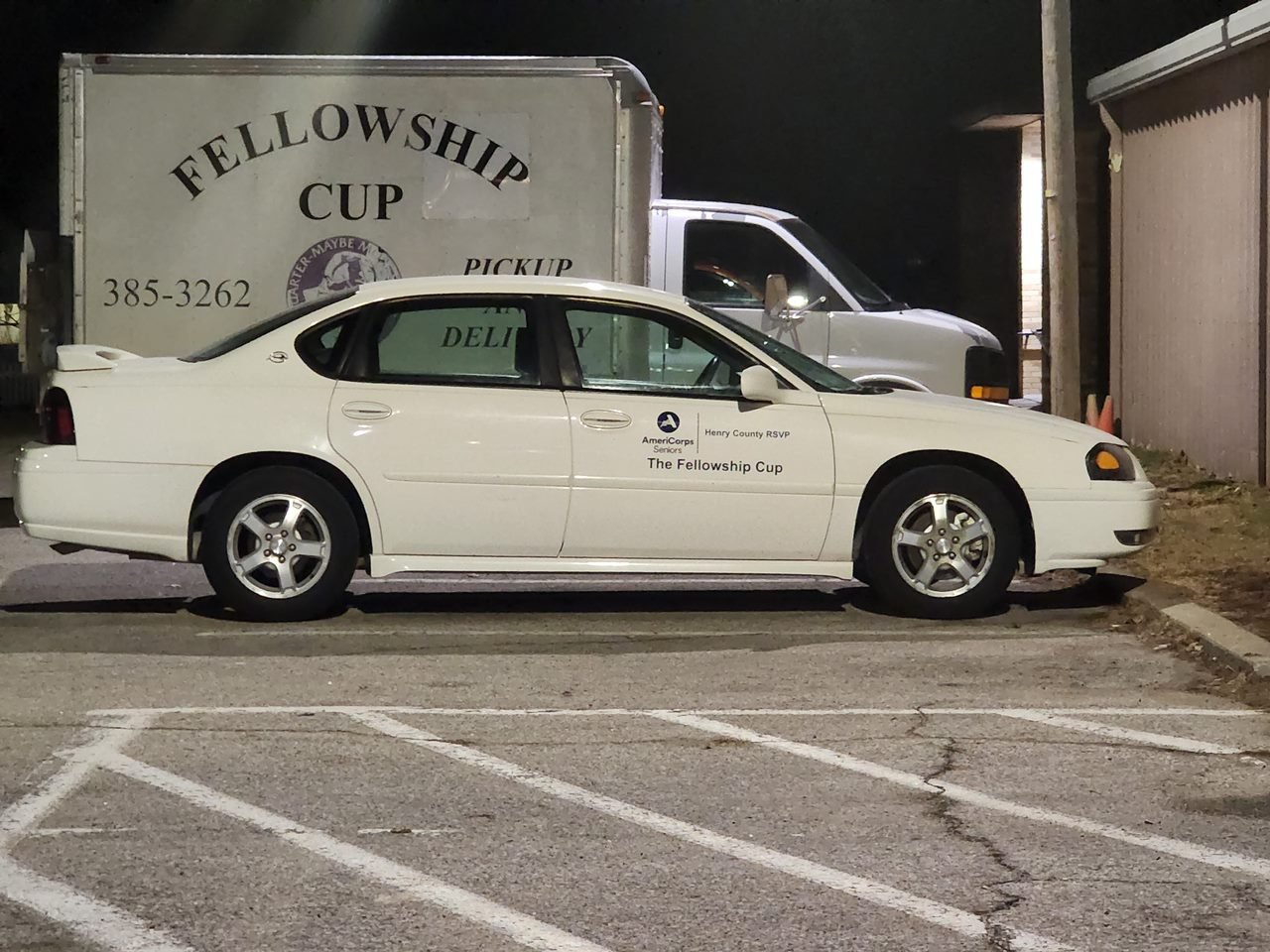 A white sedan parked in a parking lot at night, with a mobile food truck behind it. The car has stickers indicating it is associated with the Fellowship Cup and Henry County RSVP.