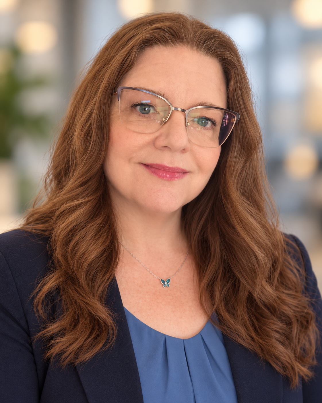 A woman with long red hair, glasses, and a butterfly necklace, smiling in an office setting.