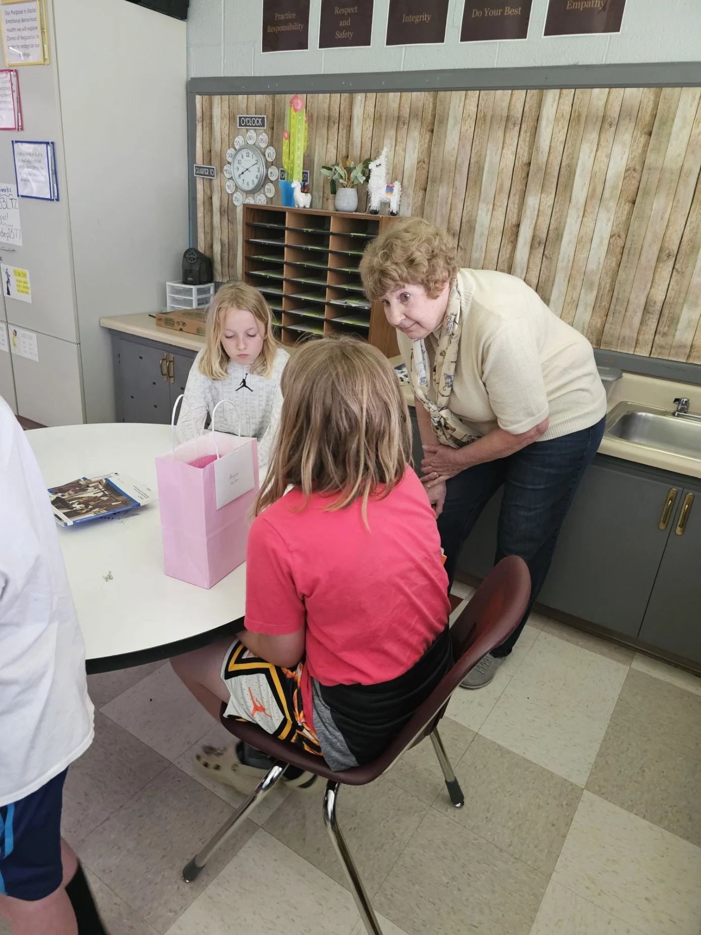 A classroom scene where a teacher is leaning over a group of students sitting at a round table. The students have their backs to the camera, and one girl with blonde hair is facing forward. There are various classroom decorations and organizational items, including a wall clock, a small shelf with green trays, and posters with motivational words like 'Practice Responsibility' and 'Respect and Safety' on the wall.