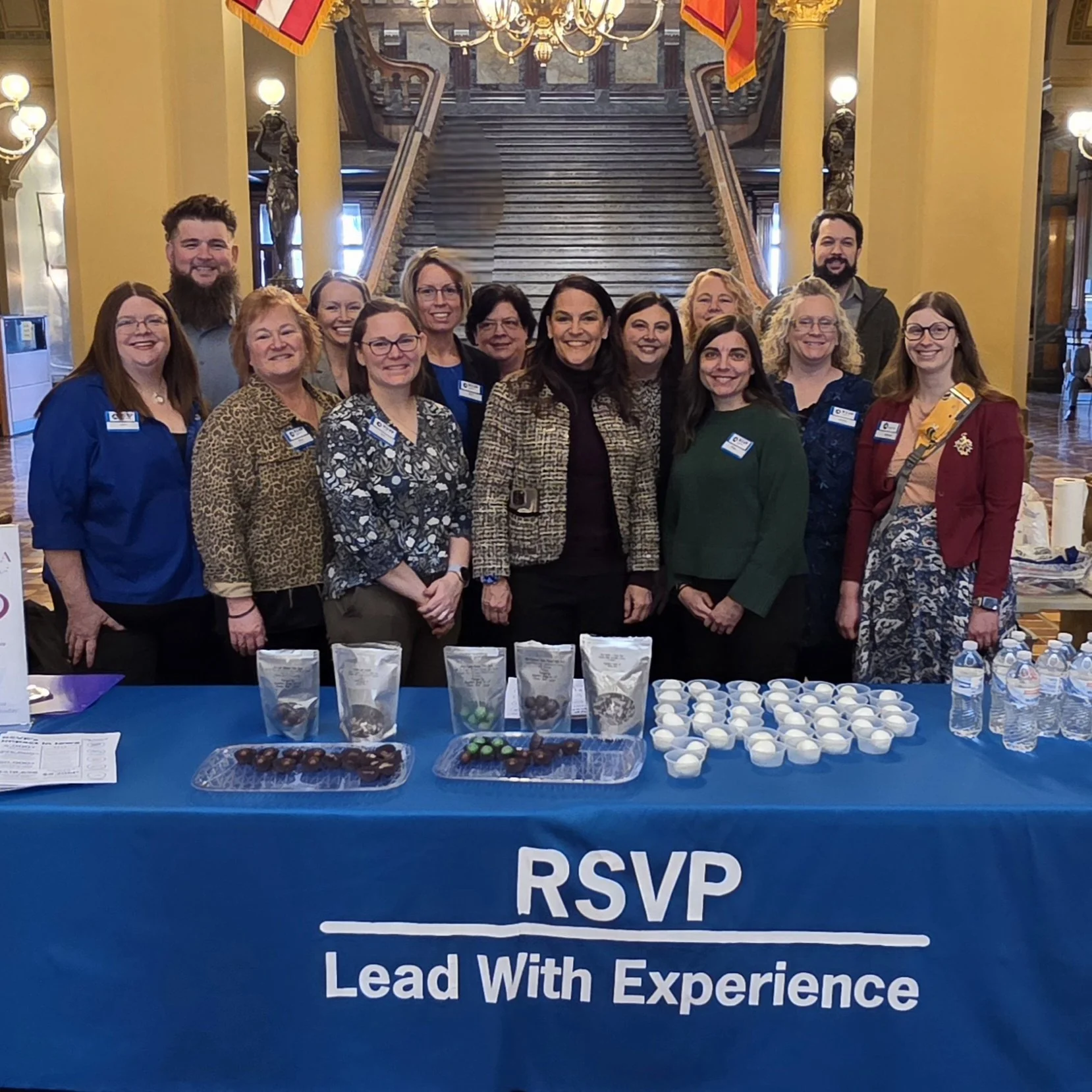 Group of fifteen people, including men and women, standing behind a table with snacks and RSVP sign at a formal indoor event with grand staircase andFlag banners in the background.