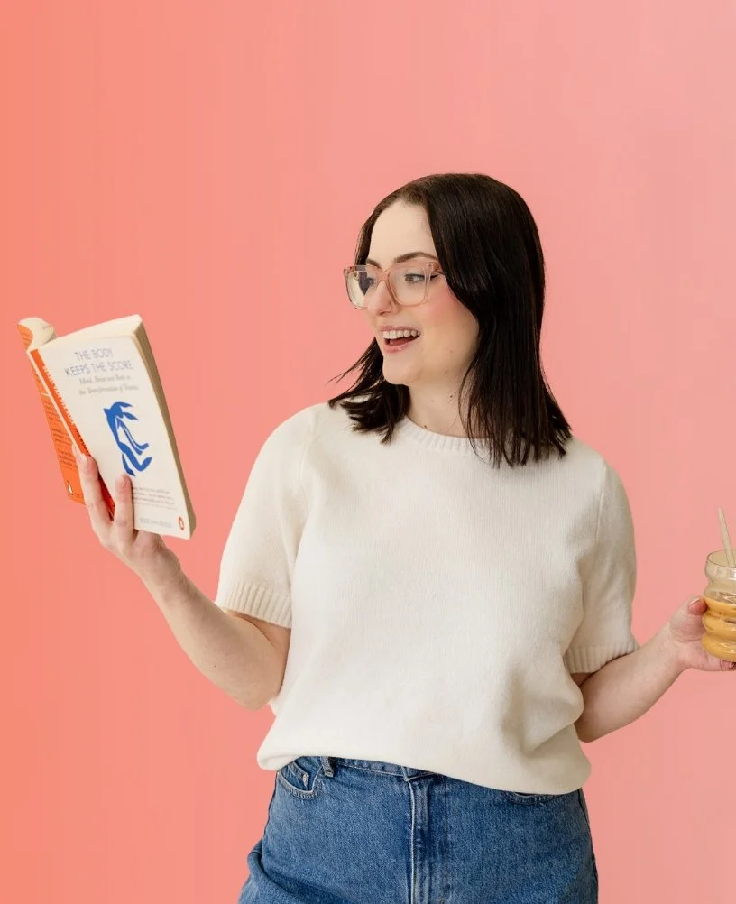 Woman with glasses holding a book and a jar of iced coffee against a pink background.