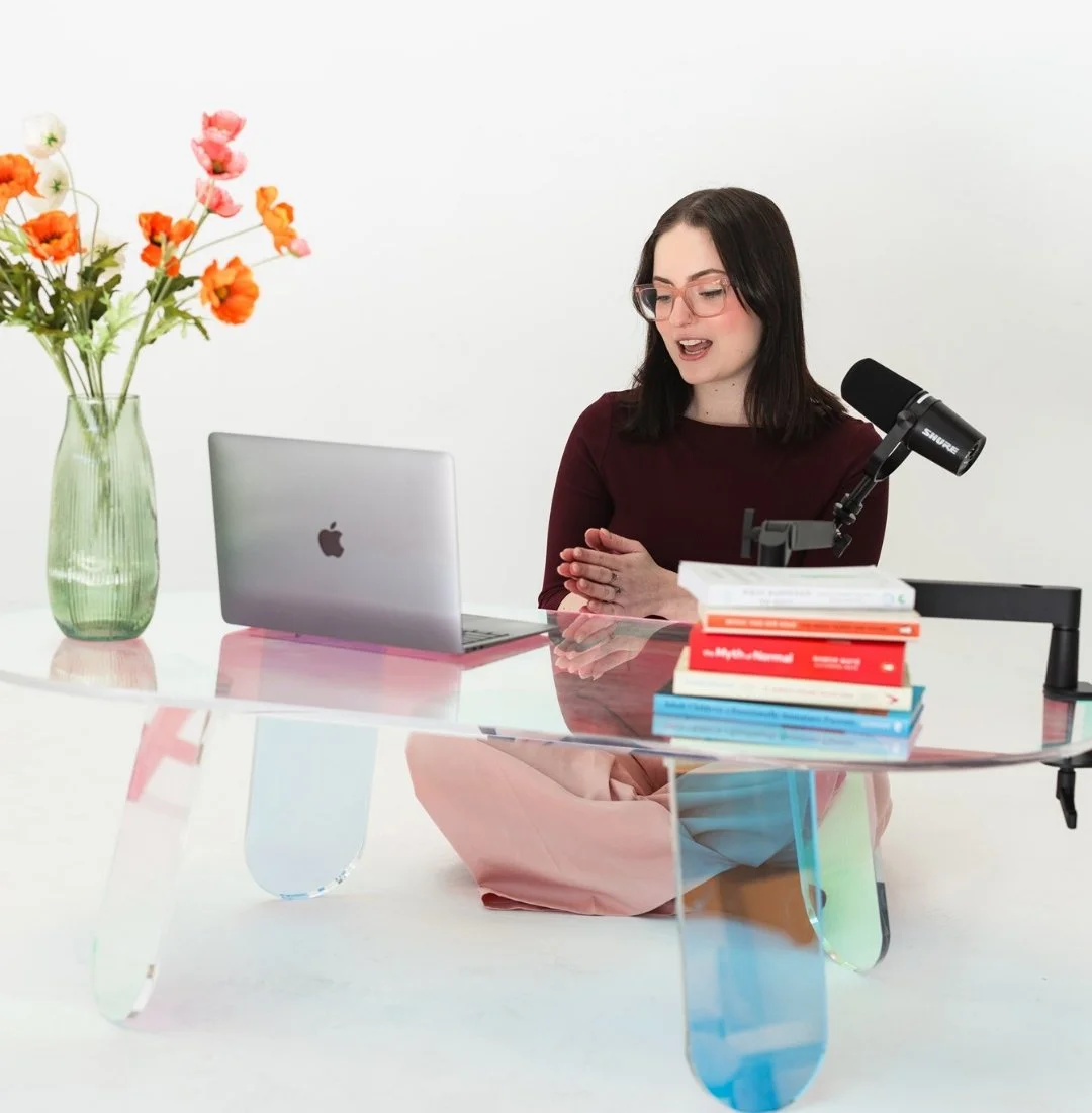 A woman with dark hair and glasses sitting cross-legged at a glass table, speaking into a microphone. On the table are a laptop, a stack of books, and a green vase with colorful flowers. The background is plain white.